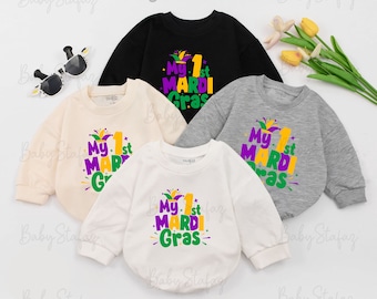 May include: Four baby onesies in different colors with the text "My 1st Mardi Gras" in purple, yellow, and green. The onesies are on a white background.