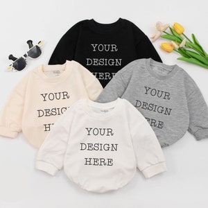 May include: Four long-sleeved baby onesies in different colors: black, beige, white, and gray. Each onesie has the text "YOUR DESIGN HERE" printed on the front.