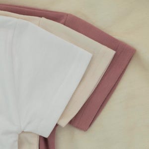 May include: A close-up of three short-sleeved t-shirts in white, beige, and mauve. The shirts are neatly stacked, showcasing the texture of the fabric and the stitching details on the sleeves.