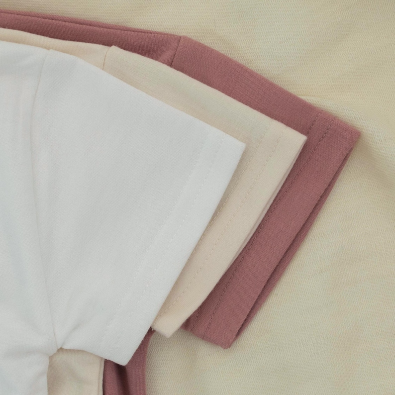 May include: A close-up of three short-sleeved t-shirts in white, beige, and mauve. The shirts are neatly stacked, showcasing the texture of the fabric and the stitching details on the sleeves.