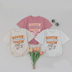 May include: Three baby bodysuits in cream, white, and mauve, each with a short-sleeved design. The bodysuits feature the text ".ROOTIN' and most definitely TOOTIN'" with a cowboy riding a horse graphic. A pair of sunglasses and flowers are also in the image.