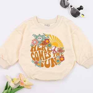 May include: A cream coloured long-sleeved baby onesie with a retro-style graphic of a sun, flowers, and peace sign. The text "Here Comes The Sun" is printed in orange and brown letters.
