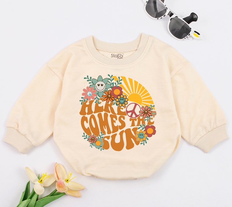May include: A cream coloured long-sleeved baby onesie with a retro-style graphic of a sun, flowers, and peace sign. The text "Here Comes The Sun" is printed in orange and brown letters.