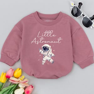 May include: A pink long-sleeved sweatshirt with the text "Little Astronaut" and a cartoon astronaut in a white spacesuit.