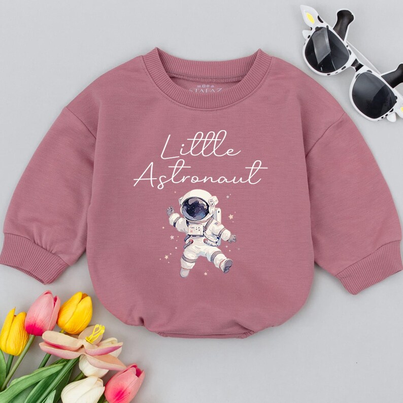 May include: A pink long-sleeved sweatshirt with the text "Little Astronaut" and a cartoon astronaut in a white spacesuit.
