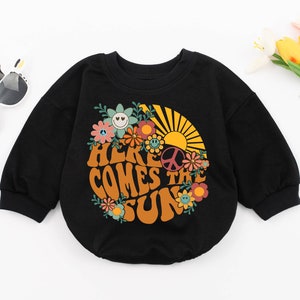 May include: Black long-sleeved sweatshirt with a retro-style graphic print of a sun, flowers, and the text "Here Comes The Sun".