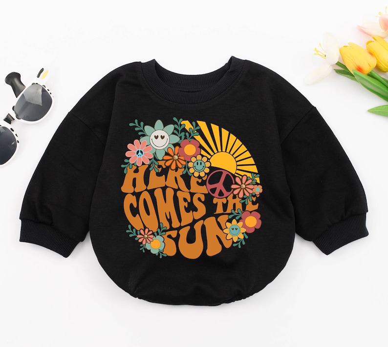 May include: Black long-sleeved sweatshirt with a retro-style graphic print of a sun, flowers, and the text "Here Comes The Sun".