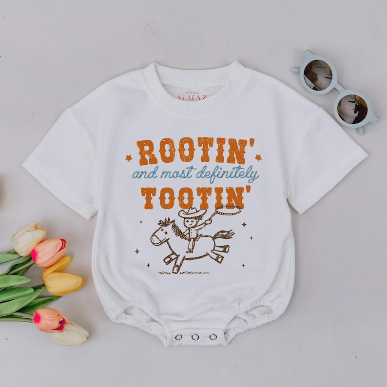 May include: White baby onesie with short sleeves and a round neckline. The onesie features a graphic of a cowboy riding a horse with the text "Rootin' and most definitely Tootin'" in orange and blue.