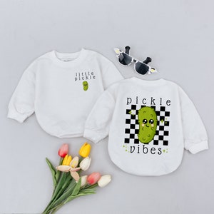 May include: Two white long-sleeved shirts with a green pickle graphic. One shirt says "little pickle" and the other says "pickle vibes" with a black and white checkered background.