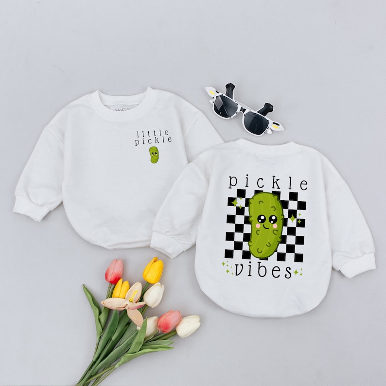May include: Two white long-sleeved shirts with a green pickle graphic. One shirt says "little pickle" and the other says "pickle vibes" with a black and white checkered background.