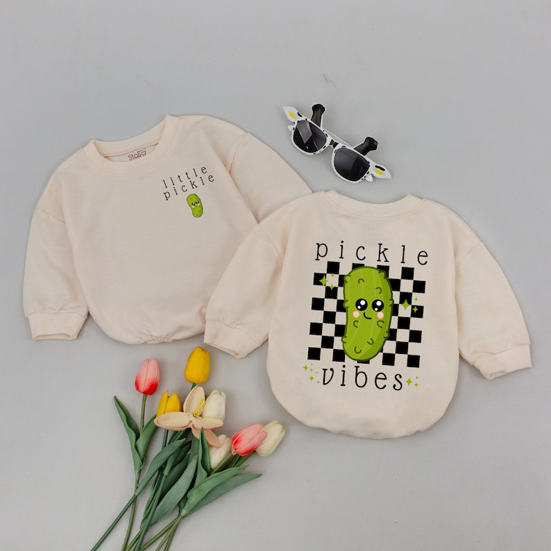 May include: Two cream colored long-sleeved shirts with a cartoon pickle graphic. One shirt says "little pickle" and the other says "pickle vibes" with a checkered background.