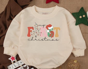 May include: A cream-colored long-sleeved onesie with the text "my first christmas" in colorful letters. A Christmas tree with lights and a Santa hat are incorporated into the letters.