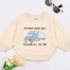 May include: Cream-colored baby onesie with the text "I'M PROOF DADDY ISN'T TRUCKING ALL THE TIME" and a blue truck graphic. The long-sleeved onesie has a crew neck and is made of soft material.