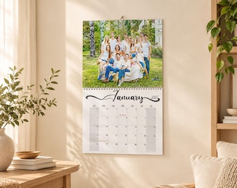 May include: A wall calendar for January 2025 featuring a large color photograph of a group of people posing outdoors on a grassy lawn. The calendar has a white background with black text and a grid layout for the days of the month, bound by a silver wire coil.