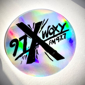 May include: A holographic sticker with a black and white graphic of a cross with the text "97X WOXY FM 97.7" on it.