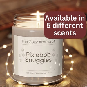 Puede incluir: Una vela encendida en un tarro de cristal con el texto "The Cozy Aroma of Pixiebob Snuggles" y "Available in 5 different scents." La vela está sobre un posavasos de madera, rodeada de luces de hadas. La vela es de 255 gramos.
