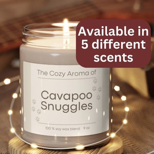 Puede incluir: Una vela encendida en un tarro de cristal con el texto "The Cozy Aroma of Cavapoo Snuggles" y "Disponible en 5 aromas diferentes". La vela está sobre un posavasos de madera, rodeada de luces de cadena. Cera de soja 100%, 255g.
