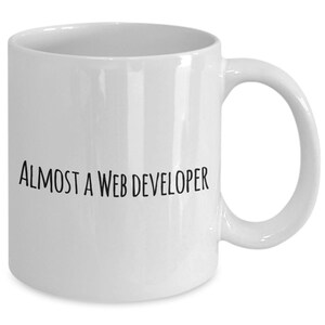 Puede incluir: Taza de cerámica blanca con asa curvada. La taza tiene el texto "ALMOST A WEB DEVELOPER" impreso en negro, con una fuente estilizada. La taza está diseñada para bebidas calientes y es adecuada para uso doméstico u oficina.