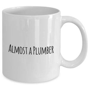 Puede incluir: Taza de cerámica blanca con el texto "ALMOST A PLUMBER" en letras negras. La taza tiene un asa curva y un acabado liso y brillante. Un regalo humorístico para un fontanero.