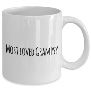 May include: A white ceramic mug with a curved handle. The mug features the text "MOST LOVED GRAMPSY" in black, handwritten-style lettering. The mug is designed for hot beverages.