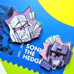 May include: Two cartoon character pins featuring Transformers characters Megatron and Starscream. Megatron is purple and grey with red eyes. Starscream is purple and grey with pink accents and red eyes. Both characters have a menacing expression.