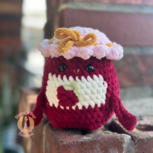 May include: A crocheted plush toy in the shape of a bird wearing a pink hat with a gold bow. The bird is holding a green leaf in its right wing. The toy is made with red, white, and green yarn.