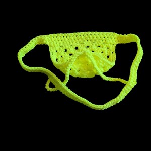May include: A neon yellow crocheted garment with a pouch and straps. The handmade item is displayed against a black background. The openwork design of the pouch is visible.