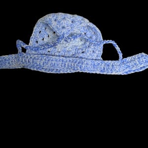 May include: A crocheted hat in shades of light blue and white, featuring a wide brim and a rounded crown. The hat has a textured appearance due to the crochet stitches and includes two straps. The hat is set against a black background.