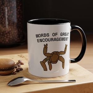 Peut inclure: Un mug en céramique blanche avec un intérieur et une anse noirs. L'avant présente le texte noir "WORDS OF GREAT ENCOURAGEMENT" au-dessus d'une illustration brune stylisée d'une créature aux membres multiples levés en signe de fête.