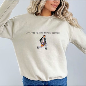 以下が含まれることがあります： ベージュのトレーナーに、たくさんの服を着た男性のグラフィックと「Could I be wearing anymore clothes?」というテキストがプリントされています