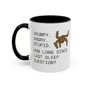 May include: A white ceramic mug with a black handle and interior. The front features a brown, multi-legged creature illustration and the black text: GRUMPY. ANGRY. STUPID. HOW LONG SINCE LAST SLEEP QUESTION? The mug has a smooth, cylindrical body.