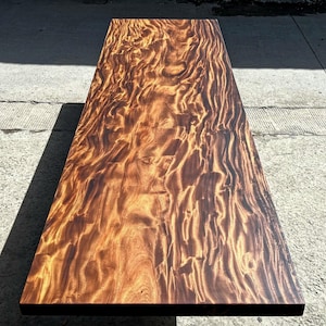 May include: A rectangular wooden table with a rich, swirling grain pattern in shades of brown and gold. The table has a smooth, polished surface and a dark edge. The wood grain creates a unique, natural design.