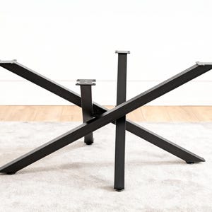 May include: Black metal table base with a modern, geometric design. The base features six angled legs that intersect at the center, creating a starburst pattern. The legs are made of thick, rectangular metal bars.