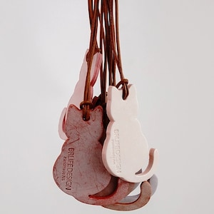 May include: A set of five leather cat keychains with brown leather cords. The keychains are in the shape of a cat and are pink and white. The keychains have the text "BRLIFEDESIGN Handmade Art" printed on them.