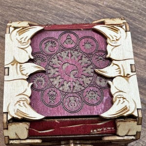 K&ouml;nnte beinhalten: Eine Holzkiste mit rotem Samt-Innenfutter und einem dekorativen Design einer Sonne mit Strahlen und anderen Symbolen. Die Kiste hat einen geschnitzten Holzrahmen mit einem dekorativen Fl&uuml;geldesign.