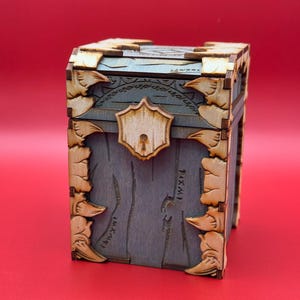 K&ouml;nnte beinhalten: Eine Holzkiste mit einem geschnitzten Design und einem Metallverschluss. Die Kiste ist dunkelblau lackiert und hat einen hellen Holzrahmen. Die Kiste steht auf einer roten Oberfl&auml;che.