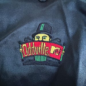 Peut inclure: Une veste en satin noir avec un écusson brodé. L'écusson représente un personnage au visage jaune portant un haut-de-forme noir, tenant une bannière rouge avec le texte Oddville et le logo MTV brodés en jaune, vert et noir.