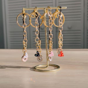 May include: A gold-colored metal display stand holding several keychains. Each keychain features a gold ring, chain, and a charm in the shape of a heart or circle. The charms are in various colors, including pink, black, and red.