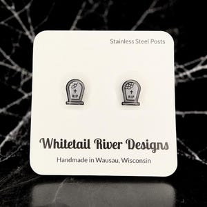 May include: A pair of silver stud earrings with a black and white tombstone design. The tombstones have a cross and the words "RIP" on them. The earrings are made with stainless steel posts. Whitetail River Designs. Handmade in Wausau, Wisconsin.