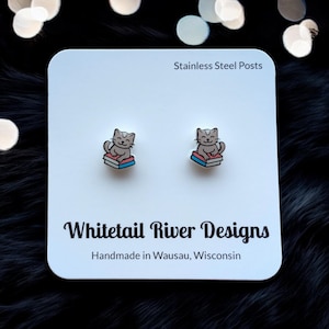 May include: A pair of stud earrings featuring a gray cat reading a book. The earrings are on a white card with the text "Stainless Steel Posts" and "Whitetail River Designs Handmade in Wausau, Wisconsin".