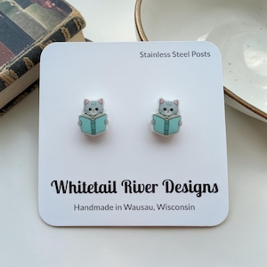 May include: A pair of cat-shaped stud earrings. Each earring features a gray cat reading a teal book. The earrings are on a white card with the text "Stainless Steel Posts" and "Whitetail River Designs Handmade in Wausau, Wisconsin."