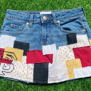 Könnte beinhalten: Blaue Jeansrock mit Patchwork-Design. Der Rock hat verschiedene Stoffflicken in Rot, Schwarz, Gelb und Weiß, einige mit gedruckten Mustern und Text. Auf einer grünen, grasbewachsenen Oberfläche platziert.