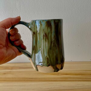 May include: A ceramic mug featuring a cylindrical body with a glossy glaze in shades of olive green, brown, and hints of blue. The glaze drips down toward the base, revealing a raw, unglazed tan clay bottom. It has a curved handle.