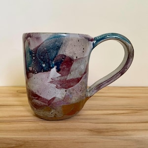 Può includere: Una tazza in ceramica con smalto maculato e multicolore caratterizzata da lavaggi astratti di blu profondo, bordeaux e bianco. La tazza presenta un manico curvo e un corpo liscio e arrotondato, con una tonalità marrone terrosa alla base.