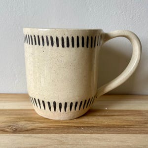 May include: Cream-colored ceramic mug with a large handle. The mug features a speckled surface and is decorated with black vertical lines near the top and bottom. The mug is sitting on a wooden surface.