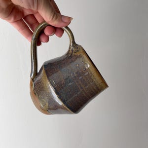 May include: A ceramic mug with a glossy, multi-toned glaze featuring deep brown, bronze, and earthy tan hues. The mug has a rounded body, a wide opening, and a curved handle attached to the side, showcasing a smooth, reflective surface finish.