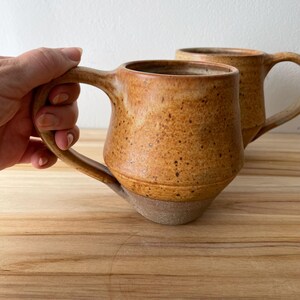 May include: Two handmade ceramic mugs featuring a speckled golden-brown glaze on the upper portion and a contrasting unglazed matte grey base. Each mug has a wide, rounded body, a sturdy handle, and a slightly tapered bottom design.