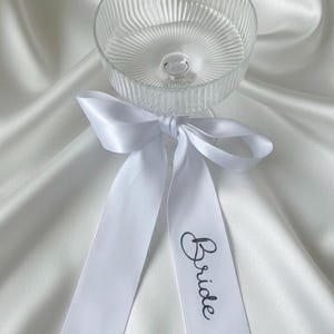 Op de afbeelding: Een heldere champagnecoupe met verticale geribbelde details, vastgebonden met een wit satijnen lint. Het lint heeft het woord "Bride" in elegant zwart schrift, perfect voor een bruiloft.