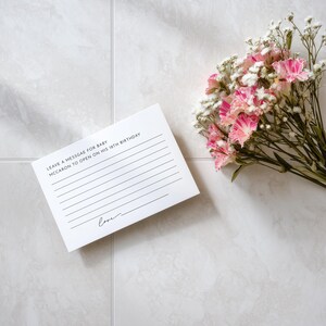 May include: A white card with lined spaces and the text "LEAVE A MESSGAE FOR BABY MCCARON TO OPEN ON HIS 18TH BIRTHDAY" and the word "love" in script. A bouquet of pink and white flowers rests nearby.