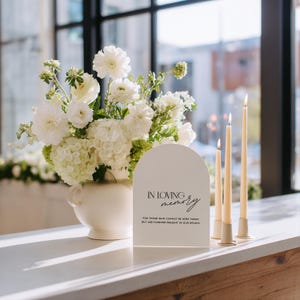 May include: A white sign with the text "IN LOVING MEMORY" is placed in front of a floral arrangement and lit candles. The sign reads "FOR THOSE WHO CANNOT BE HERE TODAY BUT ARE FOREVER PRESENT IN OUR HEARTS". The arrangement features white flowers in a white vase.